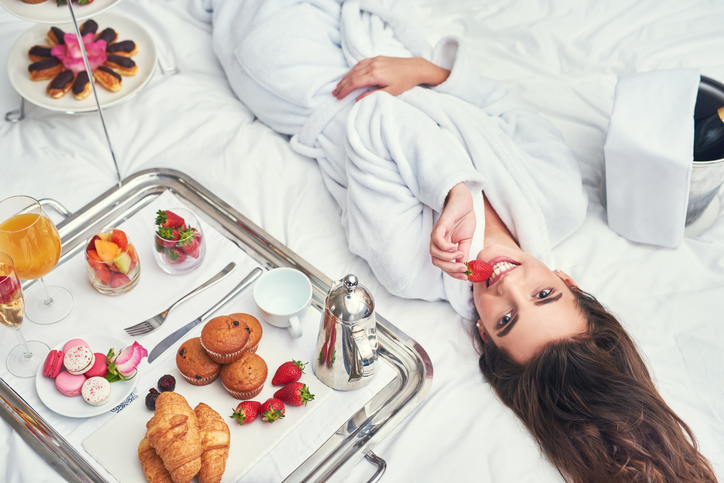 A woman lying on her bed while enjoying the desserts and wine beside her